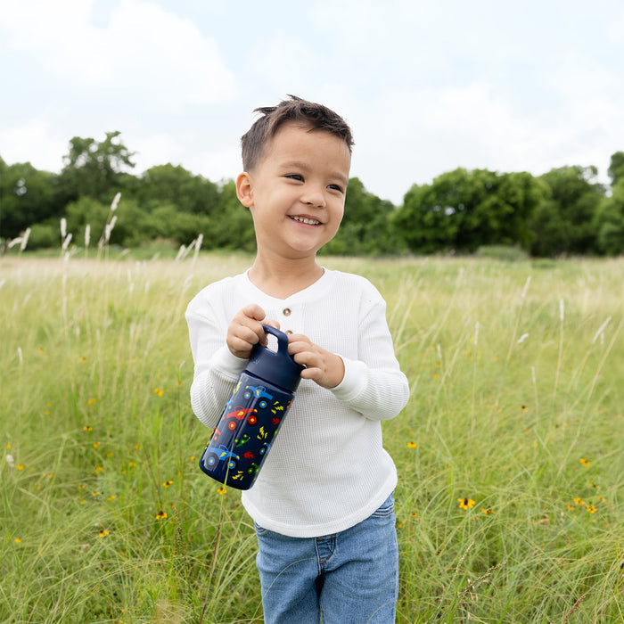 Child holding colorful navy Simple Modern water bottle with geometric patterns in outdoor setting with grass and wildflowers, wearing white long-sleeve shirt and blue jeans.