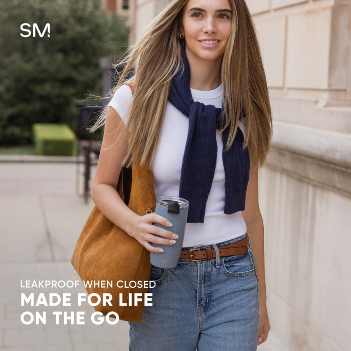 Woman in casual outfit holding gray Simple Modern tumbler with black lid in upscale urban courtyard setting. Text reads "Leakproof when closed, made for life on the go."
